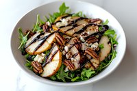 Fresh pear and pecan salad with arugula greens, topped with balsamic glaze and crumbled blue cheese on white ceramic plate.