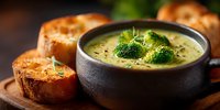 Creamy broccoli soup in black bowl with fresh herbs and crusty bread slices on wooden board for healthy comfort food dining and restaurant menu.