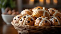 Fresh hot cross buns with raisins in woven basket, glazed with white icing cross on top, traditional Easter baking, warm brown tones, selective focus.