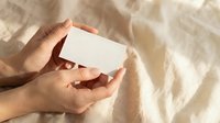 Hands holding blank white business card mockup on soft beige fabric background with natural sunlight shadows for branding design presentation.