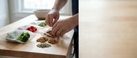 Male hands chopping nuts on wooden cutting board with fresh vegetables in containers nearby, food preparation and healthy cooking concept.