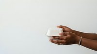 Hands of a woman with dark skin tone holding an empty white takeout container or food box against a neutral background, suitable for food delivery, restaurant, catering, and packaging concepts.