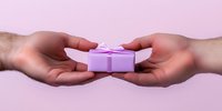 Hands exchanging small pink gift box with bow on pastel pink background. Romantic gesture showing giving and receiving present between two people.