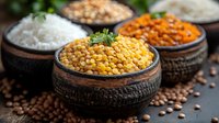 Yellow lentils, rice and spices in rustic clay bowls on dark background with scattered legumes and fresh herbs, traditional Indian cuisine setup.