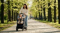Young Asian mother pushing baby stroller through tree-lined park pathway on sunny day. Family outdoor activity and parenting lifestyle concept.