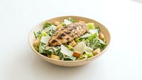 Grilled chicken Caesar salad with romaine lettuce, croutons, and shaved parmesan in a wooden bowl on white background.