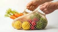 Caucasian male hands holding a reusable mesh bag filled with fresh vegetables and fruits, promoting eco-friendly sustainable shopping lifestyle.