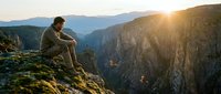 Male hiker in casual outdoor clothing sitting on rocky cliff edge overlooking vast mountain canyon valley at golden hour sunrise or sunset, contemplating nature and adventure.