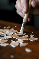Close-up of hand holding paintbrush cleaning scattered eggshell fragments on wooden surface, demonstrating delicate craftsmanship in natural light.