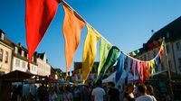 Colorful rainbow bunting flags hanging across busy town square with historic buildings and crowds celebrating outdoor festival event.