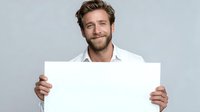 Smiling Caucasian man with beard holding blank white sign against gray background, perfect for adding custom message or promotional content.
