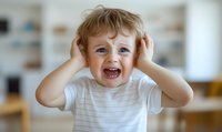 Excited Caucasian toddler boy with blue eyes wearing striped t-shirt covers ears with hands while laughing joyfully at home, showing playful expression.