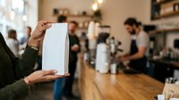 Person holding blank white box in busy cafe or coffee shop environment with staff and customers in background.