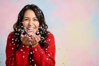 Happy young Hispanic woman in red sweater blowing colorful confetti with joyful expression against pastel rainbow background for celebration concepts.