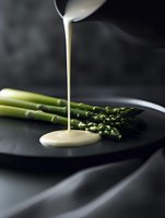 Fresh green asparagus spears on dark plate with creamy white sauce pouring from above, creating elegant food composition against black background with soft lighting.