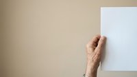 Senior adult hand holding blank white paper against neutral beige wall, suitable for mockups, presentations, and advertising campaigns.