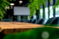 Wooden table surface with natural grain texture in modern office space, framed by blurred green fern leaves creating depth and bokeh lighting effects.