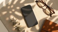 Smartphone with eyeglasses and dried eucalyptus on beige surface with natural shadow patterns from sunlight creating minimalist workspace composition.