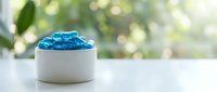 Blue hydrogel beads in white ceramic bowl on clean surface with soft natural lighting and blurred green foliage background for skincare and beauty concepts.