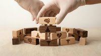 Hands in gloves carefully placing wooden block with heart symbol onto pyramid of blocks with various icons, representing care and precision in building relationships.
