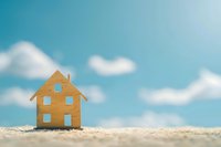 Small wooden house model on sandy beach with blue sky and white clouds background for real estate and vacation concepts.