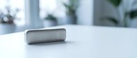 Modern silver portable wireless speaker on white desk in bright office interior with plants and natural lighting for technology presentations.