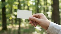 Hand holding blank white business card in natural forest setting with green bokeh background for mockup and branding purposes.