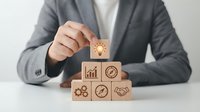 Professional businesswoman in gray suit arranging wooden blocks with business icons including lightbulb, growth chart, and gears on white desk.