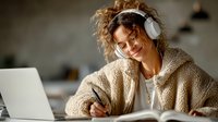 Young woman with curly hair wearing headphones and cozy sweater writing in notebook while working on laptop at home office desk for remote work and studying.