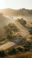 Winding rural road through golden hills with morning mist, single vehicle traveling through scenic landscape with oak trees and dry grassland during sunrise or sunset.