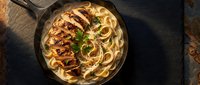Creamy pasta with grilled chicken breast and fresh herbs in cast iron skillet on rustic wooden table for restaurant menu and food photography.