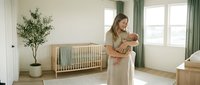 Young Caucasian mother in beige dress holding newborn baby in bright modern nursery with wooden crib and green curtains for parenting and childcare content.