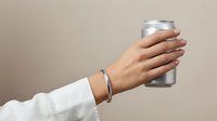 Hand holding aluminum beverage can with silver bracelet on neutral beige background for modern lifestyle and refreshment concepts.