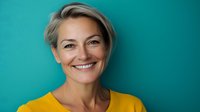 Confident middle-aged woman with short blonde hair wearing yellow top smiling genuinely against turquoise background, representing wellness, success and positive aging.