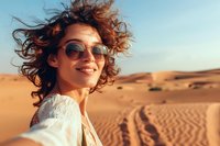 Woman with curly reddish-brown hair and round sunglasses smiling in desert landscape at golden hour. Wearing white shirt. Travel and lifestyle concept.