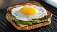 Grilled sandwich with sunny side up egg, mashed avocado, and crispy bacon on toasted bread, seasoned with black pepper, served on dark grill pan surface.