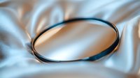 Elegant black metal bracelet on soft silky fabric with gentle folds and shadows creating minimalist jewelry composition in neutral beige tones.