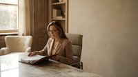 Professional businesswoman with glasses reading documents at modern office desk in warm natural lighting for corporate presentations.