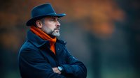 Middle-aged Caucasian man with gray beard wearing navy coat, orange scarf and blue hat in profile view against blurred autumn background with bokeh effect.