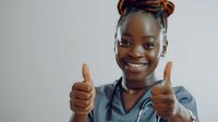 Young African American female healthcare worker in gray scrubs showing thumbs up with bright confident smile against light background. Positive medical professional portrait.