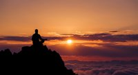 Silhouette of person meditating on mountain peak at sunrise with golden sky and sea of clouds below for mindfulness and spiritual wellness concepts.