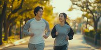 Young Asian couple jogging together in autumn park at sunset, smiling while running on tree-lined path with golden foliage background.