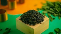 Dried green tea leaves in traditional wooden measuring box on green surface with scattered leaves and golden elements, macro food photography.