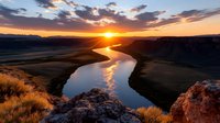 Dramatic sunset over winding river through canyon landscape with golden light reflecting on water and rocky terrain under colorful sky.