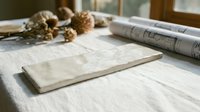 Blank marble tile sample displayed on white linen fabric with architectural blueprints and dried botanicals, showcasing interior design materials and home renovation planning concepts.