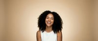 Happy young African American woman with curly hair smiling broadly in white sleeveless top against warm beige background for lifestyle and portrait photography.