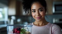 Young African American woman smiling while holding blender container with fresh fruits and berries for making healthy smoothie in modern kitchen. Wellness and healthy eating concept.