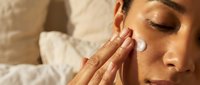 Young woman applying white facial cream moisturizer to cheek with fingers in natural sunlight for daily skincare routine and anti-aging treatment.