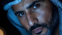 Close up portrait of middle eastern man in traditional keffiyeh headscarf with dramatic blue lighting and intense gaze against dark background.