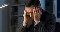Stressed businessman holding head in hands suffering from headache at office desk during late night work session.